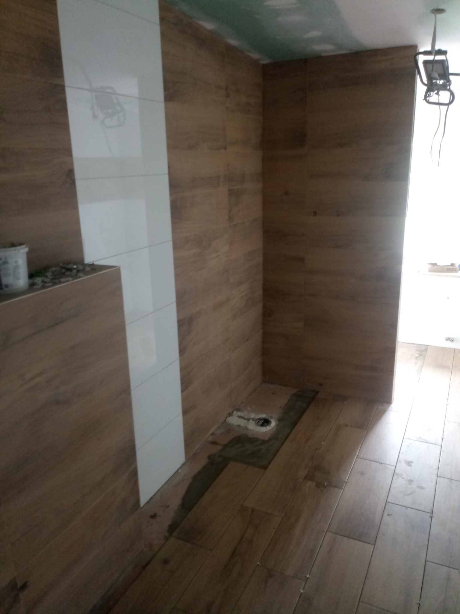 Wnętrze łazienki w trakcie wykańczania: drewnopodobne płytki na ścianach i podłodze, pionowy pas białych płytek, widoczne niedociągnięcia przy odpływie podłogowym, surowy sufit z płyt...