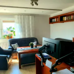 Biuro domowe z dwoma monitorami Iiyama, dokumentami, czarną sofą i fotelem, drewnianym stołem i szafką wiszącą z książkami.