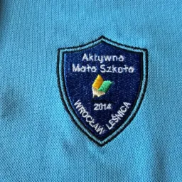 Jasnoniebieska koszulka polo z wyhaftowanym herbem 'Aktywna Mała Szkoła Wrocław Leśnica 2014' w kształcie tarczy.