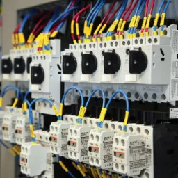 Szczegółowe ujęcie rozdzielnicy elektrycznej z bezpiecznikami, okablowaniem oznaczonym żółtymi tulejkami i precyzyjnie ułożonymi przewodami w kolorach niebieskim i czerwonym.