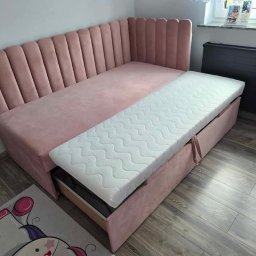 Sypialnia Snu Pablitto Paweł Kasprzak - Różowa, tapicerowana sofa narożna z wysuwanym materacem na tle drewnianej podłogi i grzejnika. Nowoczesny design z pionowymi przeszyciami na oparciu.