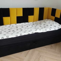 Sypialnia Snu Pablitto Paweł Kasprzak - Tapicerowany narożnik z wezgłowiem w czarno-żółte pasy, przykryty pościelą w piłki nożne. Widoczna drewniana podłoga i fragment ściany z gniazdkiem.