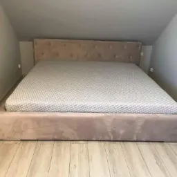 Beżowe łóżko tapicerowane z pikowanym zagłówkiem i materacem w pokoju na poddaszu z drewnianą podłogą.