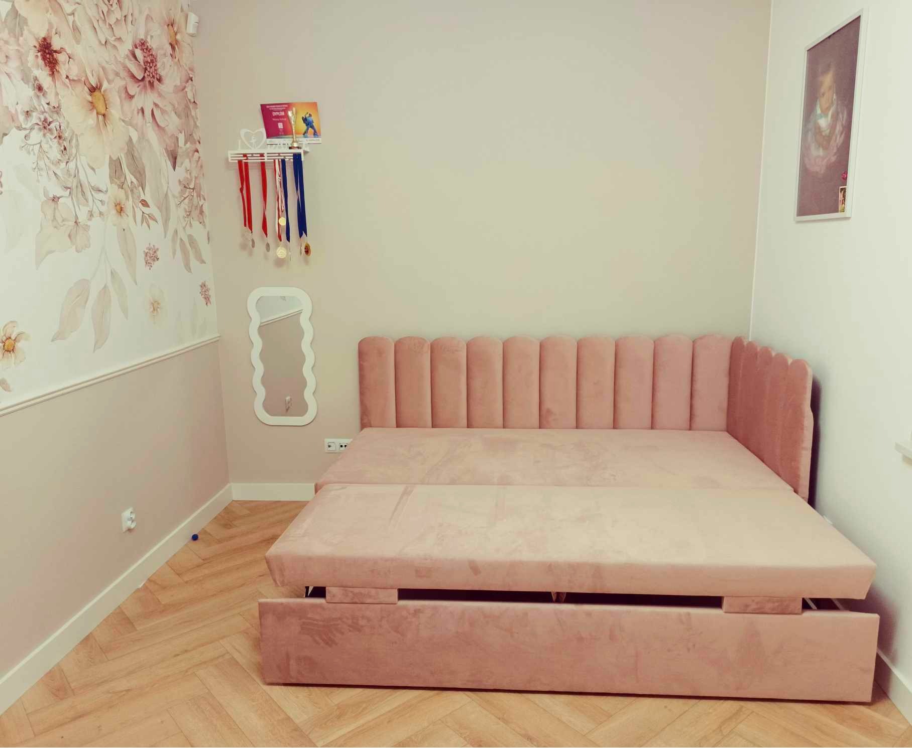 Różowe, welurowe łóżko z wysuwanym elementem w pokoju z dekoracyjną tapetą w kwiaty i półką z medalami. Widok na rozłożony mebel.