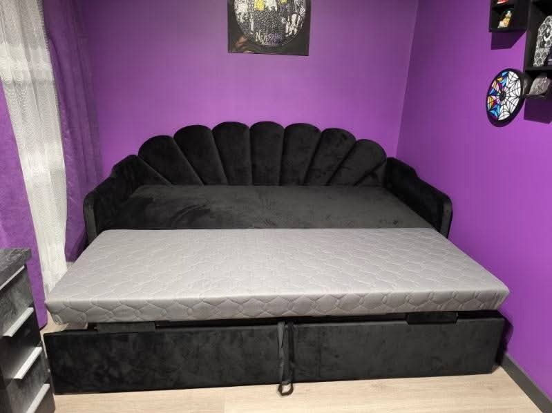 Czarna, welurowa sofa rozkładana z szarym materacem na tle fioletowej ściany z dekoracjami. Sofa z charakterystycznym, muszelkowym zagłówkiem.