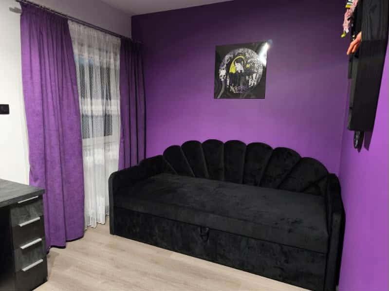 Czarna, welurowa sofa w pokoju z fioletowymi ścianami i zasłonami. Sofa z dekoracyjnym oparciem w kształcie muszli. Nad sofą wisi plakat.