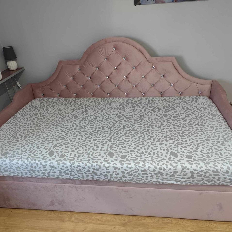 Różowe, tapicerowane łóżko z pikowanym zagłówkiem zdobionym kryształkami, pokryte prześcieradłem w panterkę, na drewnianej podłodze.