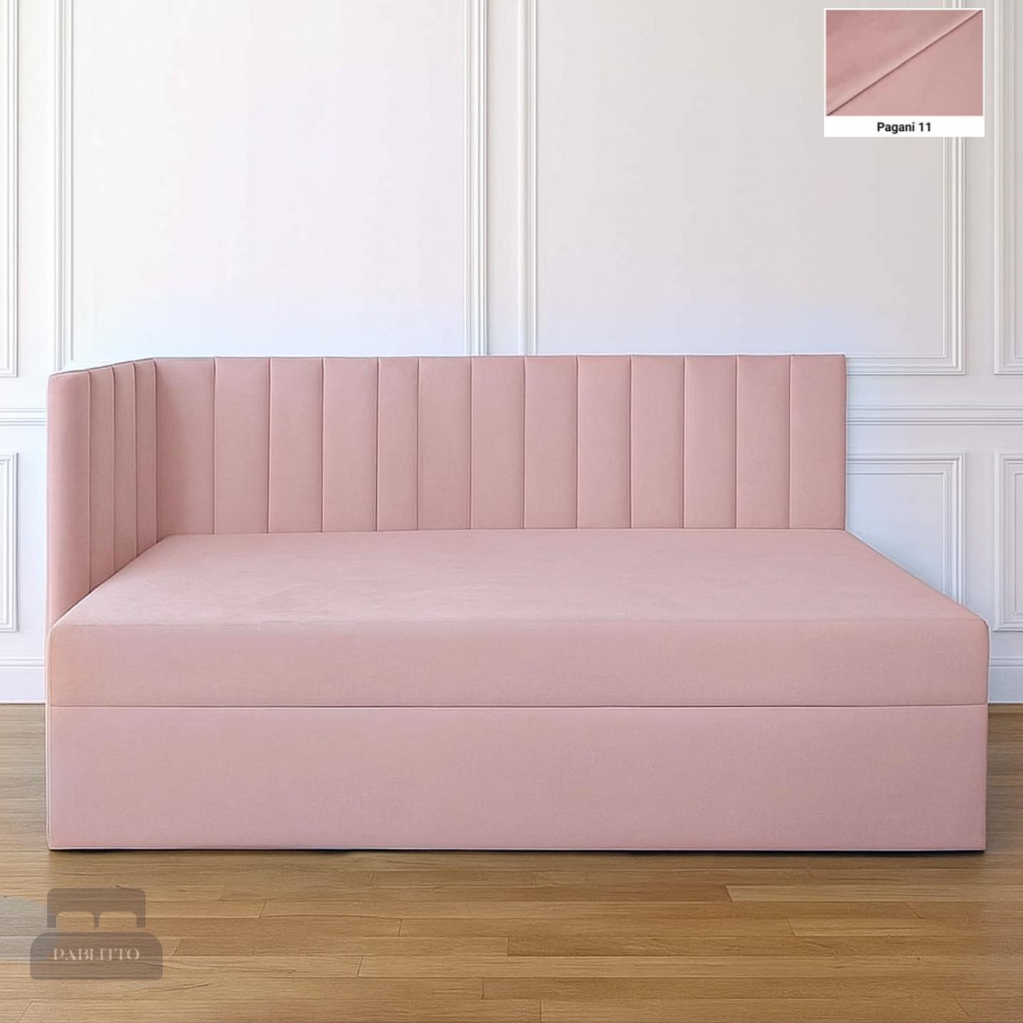 Tapicerowany, pudrowo-różowy narożnik z wezgłowiem w pionowe pasy, na drewnianej podłodze. W tle biała ściana z dekoracyjnymi listwami. W rogu próbka materiału 'Pagani 11'.