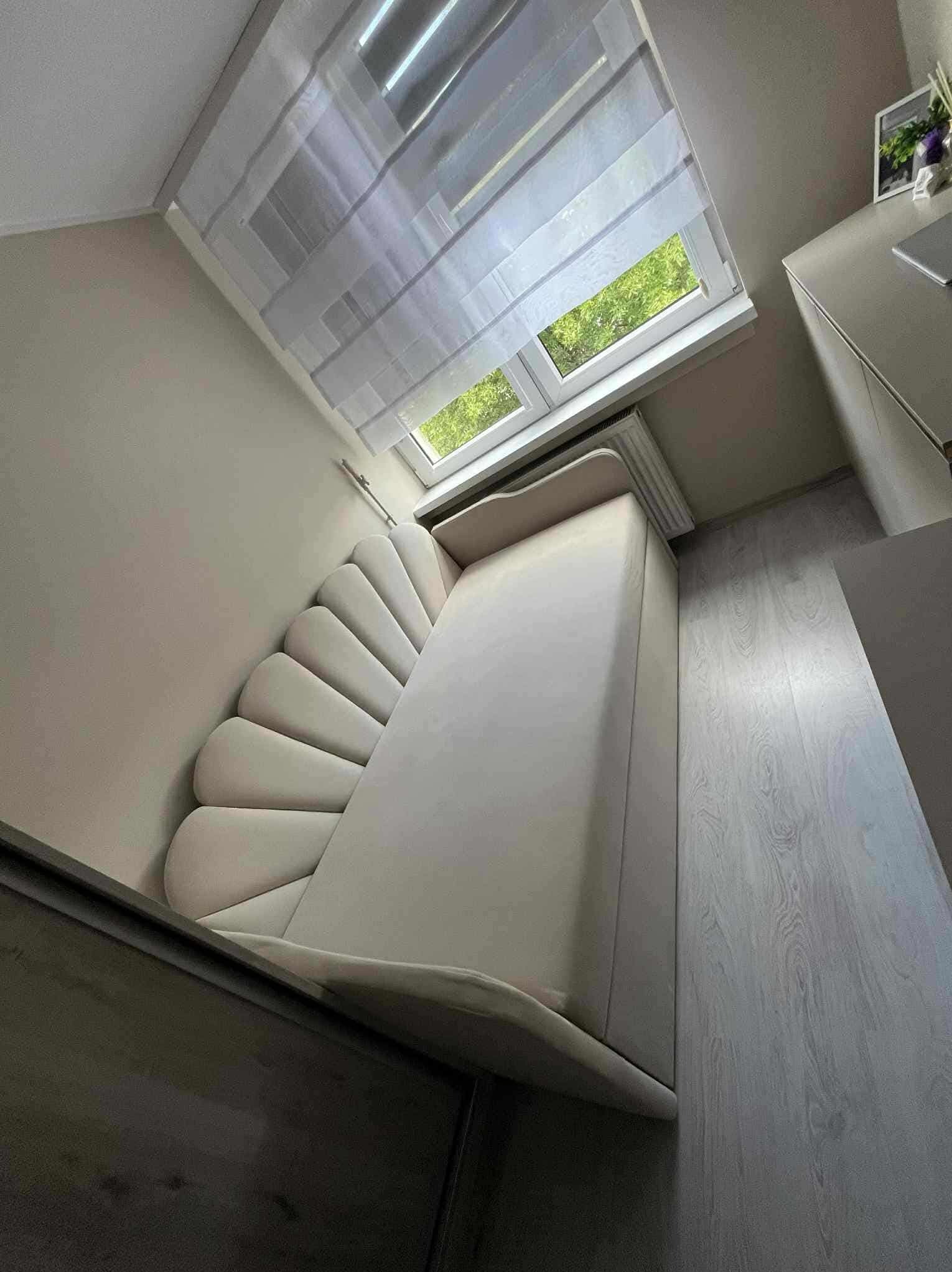 Beżowe łóżko tapicerowane z oryginalnym wezgłowiem w kształcie wachlarza, ustawione pod oknem z roletami. Minimalistyczny design w jasnym wnętrzu.