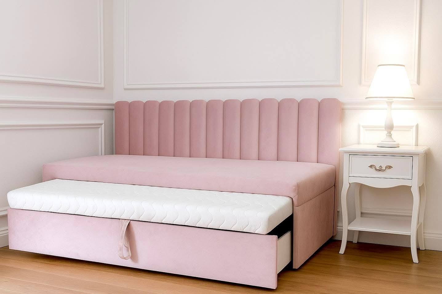 Różowa, tapicerowana sofa z wysuwanym materacem i wezgłowiem w pionowe pasy, obok biała szafka nocna z lampką, na tle ściany z dekoracyjnymi listwami.