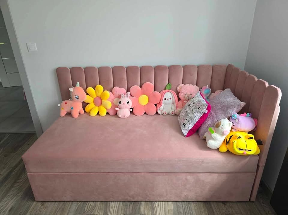 Różowa, tapicerowana sofa z pluszowymi zabawkami i poduszkami. Wezgłowie z pionowymi panelami. Ujęcie z lekkiego ukosa, ukazujące narożnik sofy.