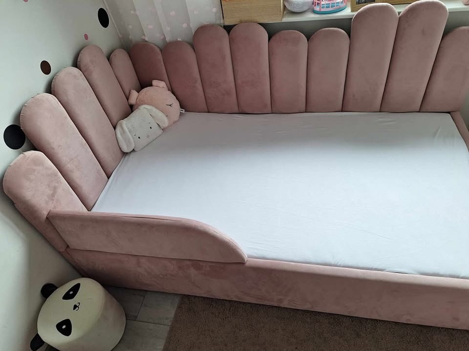 Różowe, tapicerowane łóżko dziecięce z zagłówkiem w kształcie muszelek, z pluszowymi zabawkami. Nowoczesny design, miękka tkanina, białe prześcieradło.