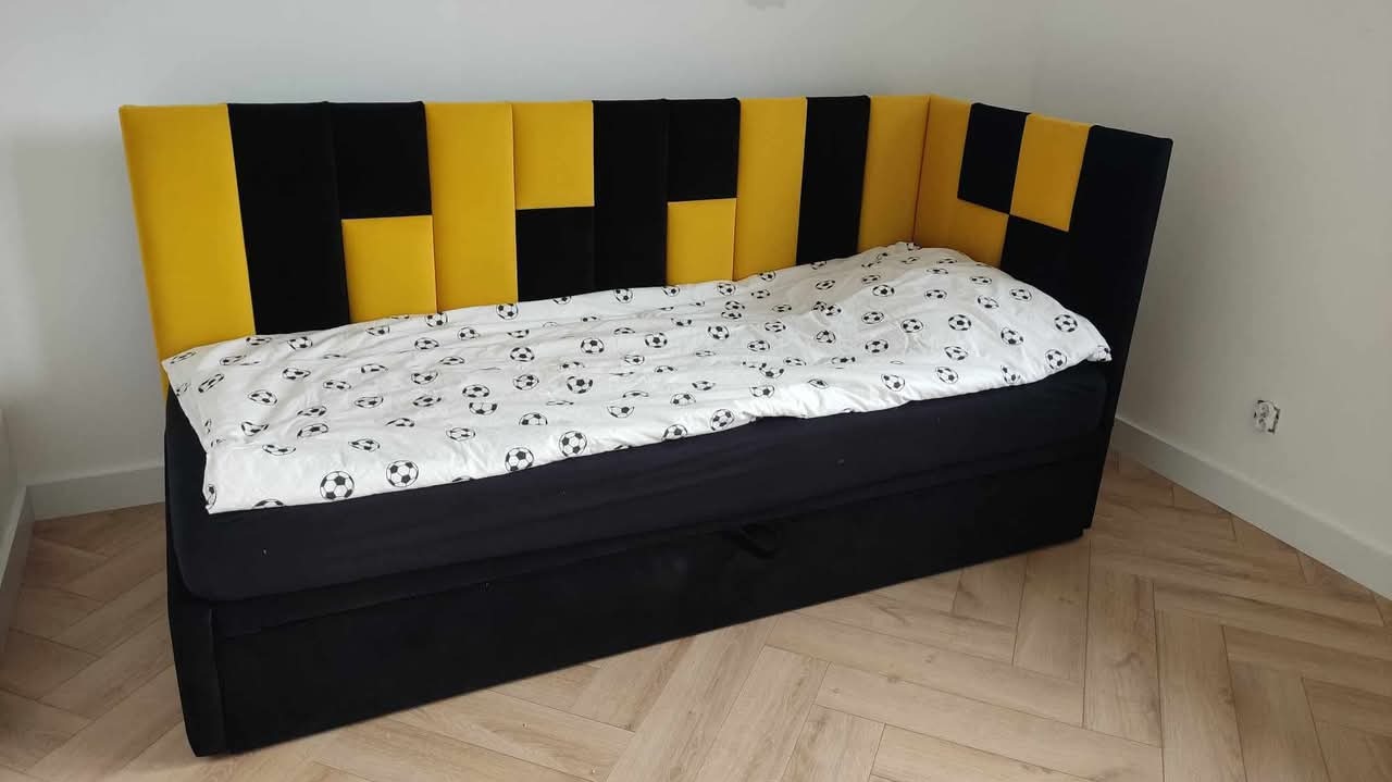 Tapicerowany narożnik z wezgłowiem w czarno-żółte pasy, przykryty pościelą w piłki nożne. Widoczna drewniana podłoga i fragment ściany z gniazdkiem.
