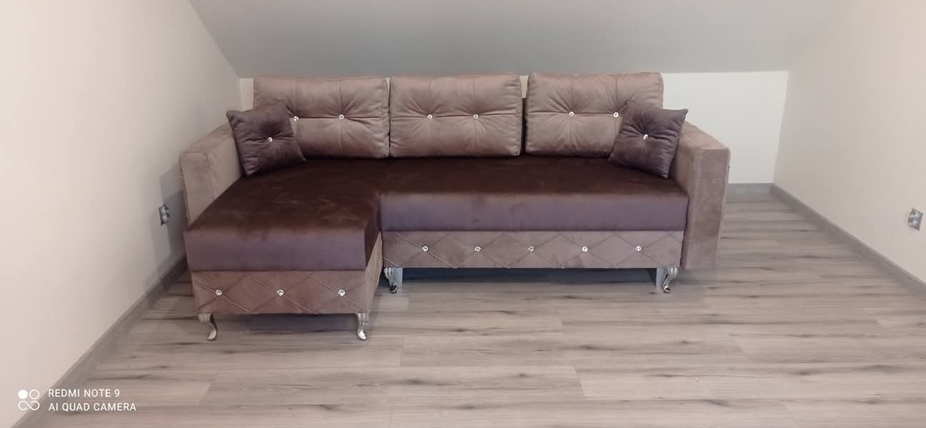 Narożnik tapicerowany w odcieniach brązu i beżu, z dekoracyjnymi przeszyciami i poduszkami, ustawiony w pokoju na poddaszu z drewnianą podłogą.