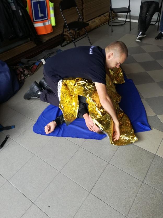 Instruktor udziela pierwszej pomocy osobie leżącej na niebieskim kocu, okrytej złotą folią termiczną; widoczny fragment wyposażenia sali szkoleniowej.