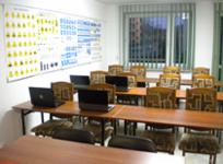 Sala wykładowa szkoły jazdy z rzędami biurek, na każdym z nich otwarty laptop, na ścianie tablica z materiałami edukacyjnymi, widok z okna na budynek.