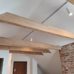 Drewniane belki stropowe w jasnym wnętrzu z ceglaną ścianą, oświetlenie punktowe na szynoprzewodzie, widoczny fragment nowo wykończonego pomieszczenia z drzwiami.