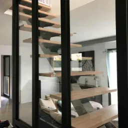 Nowoczesne, drewniane schody wiszące z czarnymi wspornikami, widoczne przez szklaną ścianę z czarną ramą w jasnym wnętrzu domu.