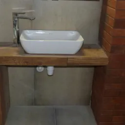 Biała, prostokątna umywalka nablatowa z chromowaną baterią, osadzona na drewnianym blacie w łazience wykończonej płytkami imitującymi cegłę.