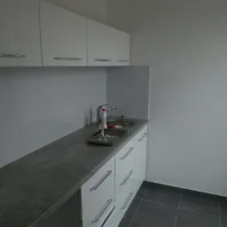 Nowoczesna łazienka z szarym blatem, białymi szafkami z podłużnymi uchwytami, chromowaną baterią i szarą podłogą z płytek.