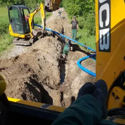 Widok z kabiny żółtej koparki JCB na wykop z ułożonymi niebieskimi rurami, z pracownikami w zielonych kombinezonach, w tle zielone drzewa i operator z nogami opartymi o deskę rozdzielczą.