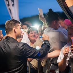 Martyna i Rafał Fotografia - Mężczyzna w czarnej kurtce rozdaje autografy na tle zgromadzonego tłumu fanów, którzy robią zdjęcia telefonami komórkowymi.
