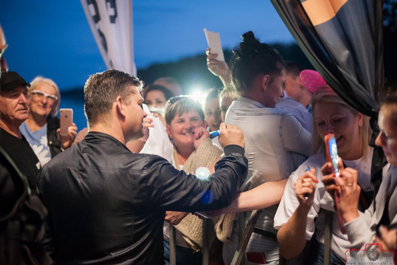 Mężczyzna w czarnej kurtce rozdaje autografy na tle zgromadzonego tłumu fanów, którzy robią zdjęcia telefonami komórkowymi.