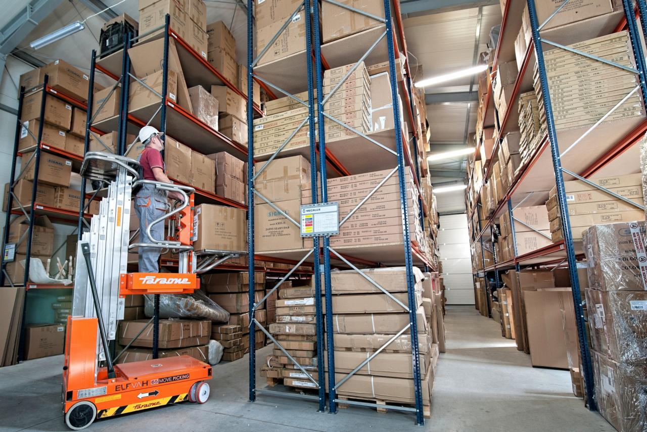 Pracownik w kasku na podnośniku Taravone ELF VAH 65 Move Picking w magazynie z regałami wypełnionymi kartonami.