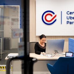 Centrum Ubezpieczeń PARTNER - Recepcjonistka uśmiecha się siedząc za biurkiem z komputerami w nowoczesnym biurze Centrum Ubezpieczeń Partner, widoczne logo firmy na ścianie.