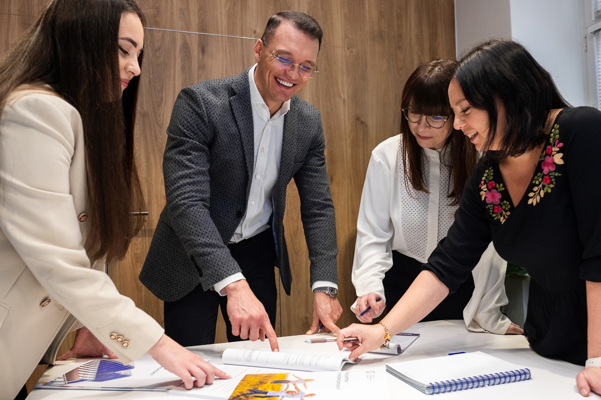 Cztery osoby analizują dokumenty na stole w biurze, mężczyzna w szarej marynarce uśmiecha się, wskazując na otwartą broszurę.