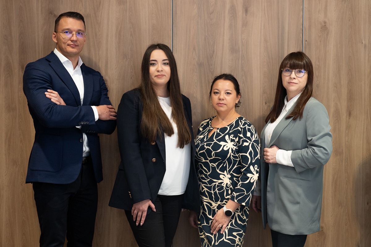 Czteroosobowy zespół w formalnym stroju pozuje na tle drewnianej ściany, prezentując profesjonalizm i zaufanie.