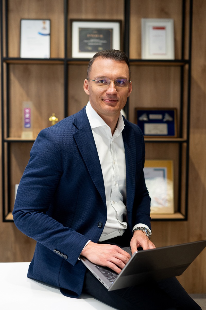 Mężczyzna w okularach i granatowej marynarce siedzi na biurku z laptopem, w tle regał z certyfikatami i nagrodami.