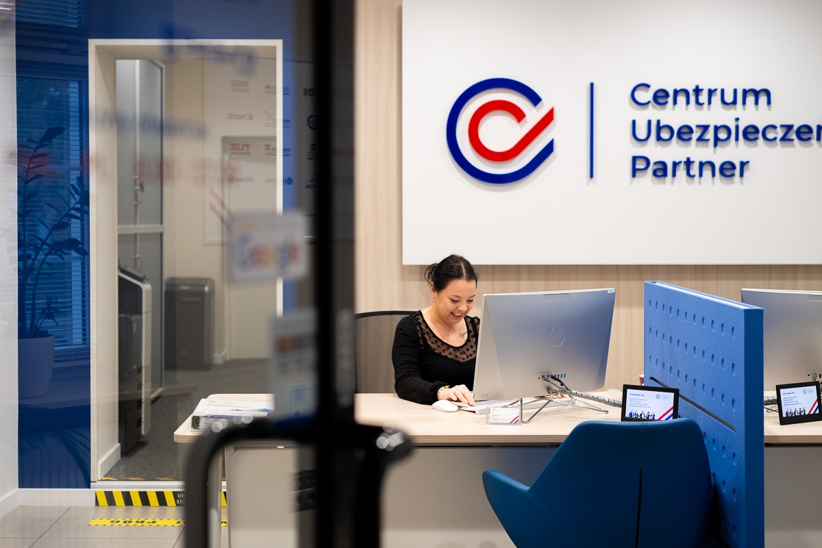 Recepcjonistka uśmiecha się siedząc za biurkiem z komputerami w nowoczesnym biurze Centrum Ubezpieczeń Partner, widoczne logo firmy na ścianie.