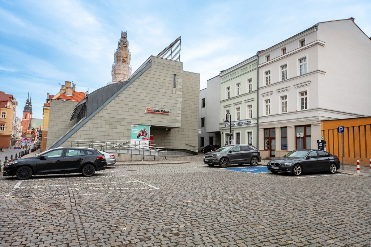 Ulica z budynkami, w tym Bank Pekao, oraz zaparkowanymi samochodami na brukowanym parkingu, w tle wieża kościoła w rusztowaniach.