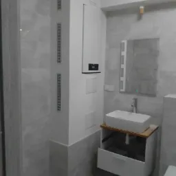 Nowoczesna łazienka z umywalką nablatową na drewnianym blacie, białą szafką z otwartą szufladą, grzejnikiem panelowym i lustrem z oświetleniem LED, wykończona szarą płytką imitującą beton.