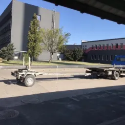 Niebieska ciężar&oacute;wka z przyczepą przewożąca długi, ciemny element konstrukcyjny na tle budynk&oacute;w przemysłowych w słoneczny dzień.