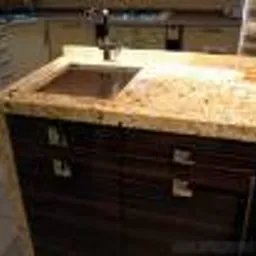 Beżowy blat granitowy z wbudowanym czarnym zlewem w nowoczesnej kuchni. Widoczne ciemnobrązowe szafki z metalowymi uchwytami i fragment jasnej ściany w tle.