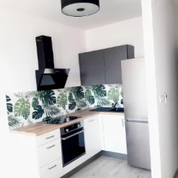 Nowoczesna, jasna kuchnia z białymi szafkami, drewnianym blatem i ścianą z motywem tropikalnych liści, czarny okap i srebrna lodówka, oświetlona okrągłą lampą sufitową.