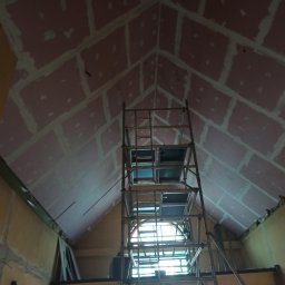 JASHAUS - Wnętrze poddasza z ukośnym sufitem pokrytym różowymi płytami z zaszpachlowanymi łączeniami. W centrum metalowe rusztowanie, w tle okno.