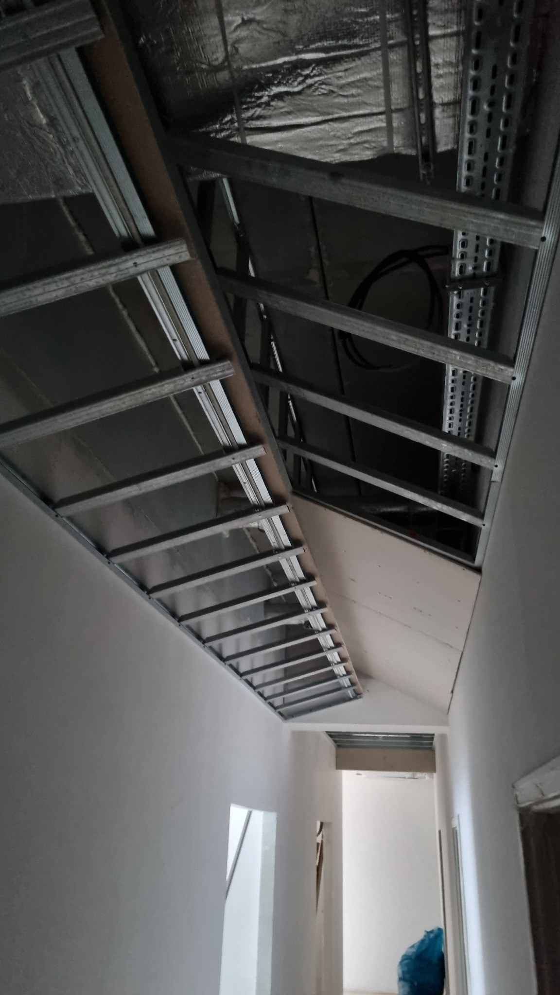 Metalowa konstrukcja sufitu podwieszanego z widoczną izolacją i przewodami w trakcie montażu w biurze, w tle widoczne kolejne pomieszczenia.