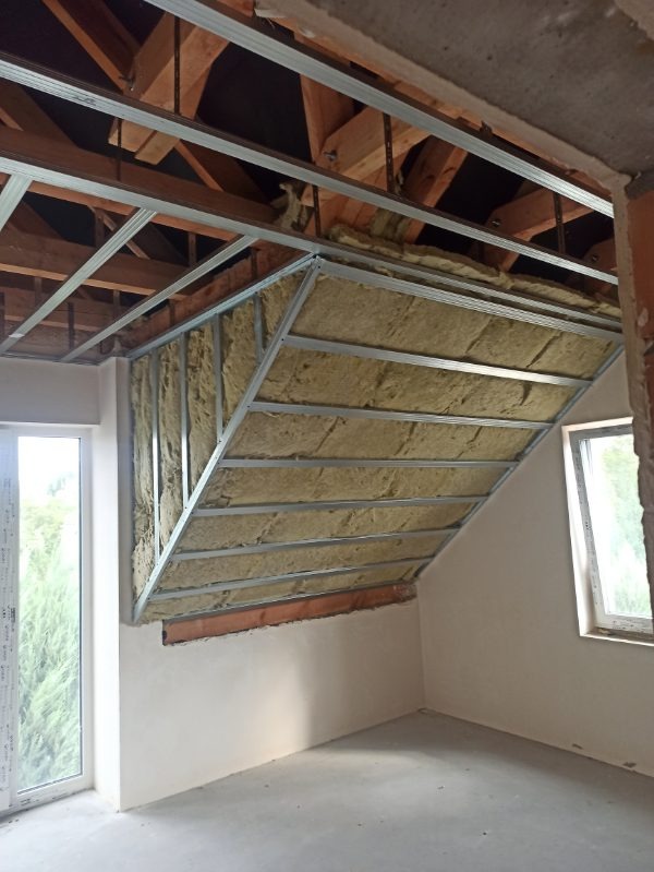 Skośny sufit poddasza w trakcie izolacji wełną mineralną, widoczne metalowe profile i drewniana konstrukcja dachu. Okno po lewej stronie kadru.