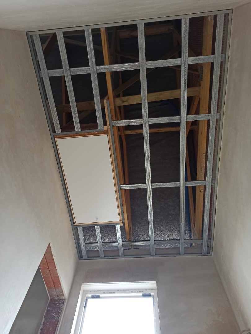 Metalowa konstrukcja sufitu podwieszanego z płyt karton-gips, widoczna od dołu, z lukiem rewizyjnym i oknem w tle. Ściany w stanie surowym.