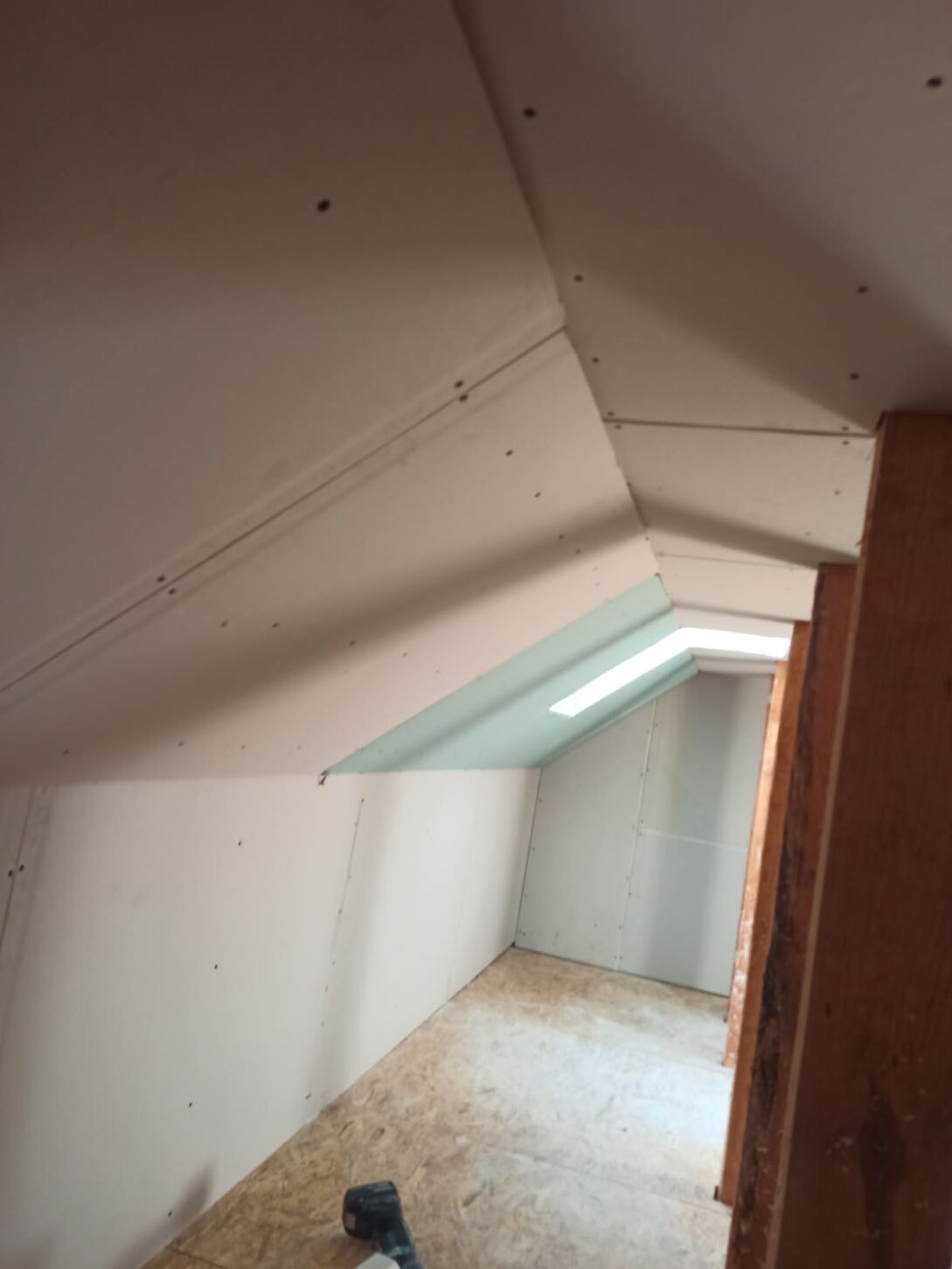 Poddasze z zamontowanymi płytami kartonowo-gipsowymi, widoczne łączenia i wkręty, surowe wykończenie, okno dachowe, podłoga z płyt OSB, narzędzie w rogu.