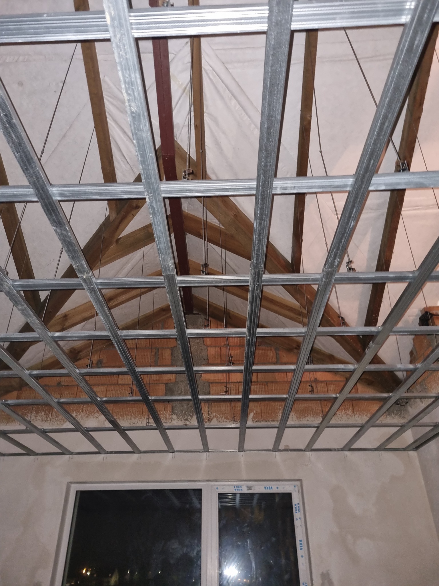 Metalowa konstrukcja sufitu podwieszanego z widoczną drewnianą więźbą dachową i fragmentem murowanej ściany, widok z dołu na okno.