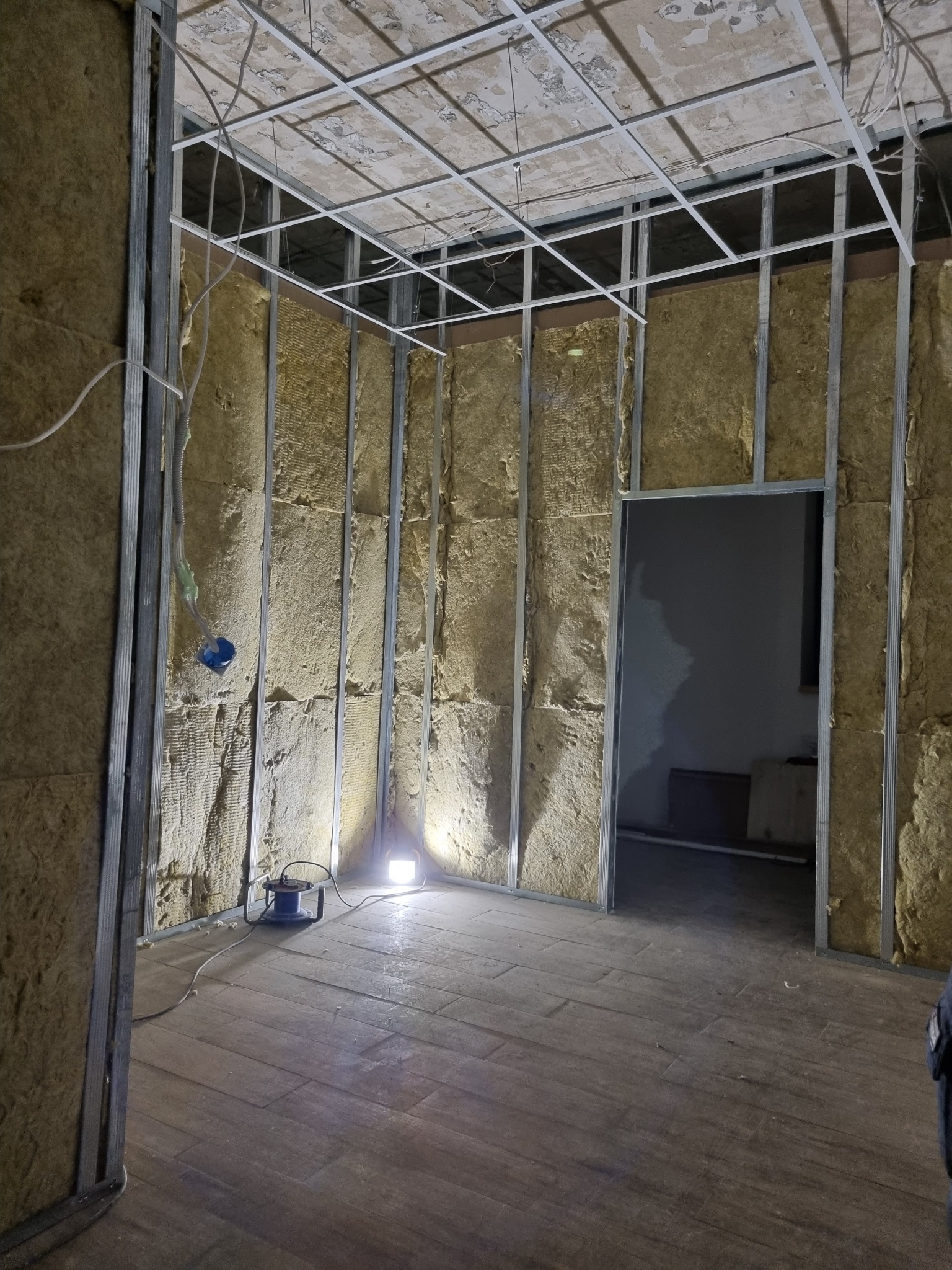 Wnętrze w trakcie prac wykończeniowych: metalowy stelaż, izolacja z wełny mineralnej, częściowo ułożona podłoga z paneli, widoczna lampa robocza i instalacja elektryczna.