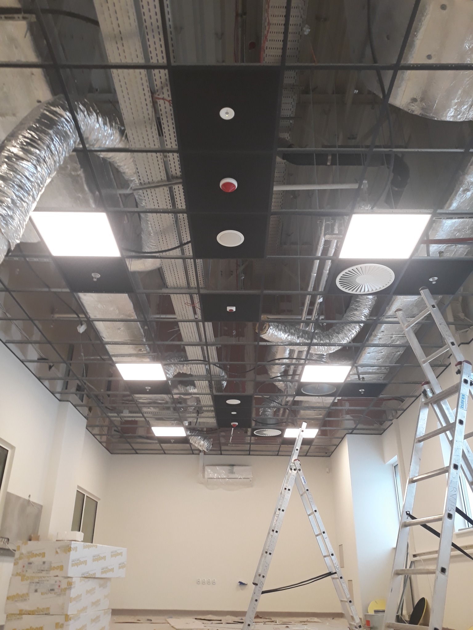 Montaż instalacji sufitowej w biurze, widoczne elementy wentylacji, oświetlenia, czujniki dymu oraz drabiny Krause