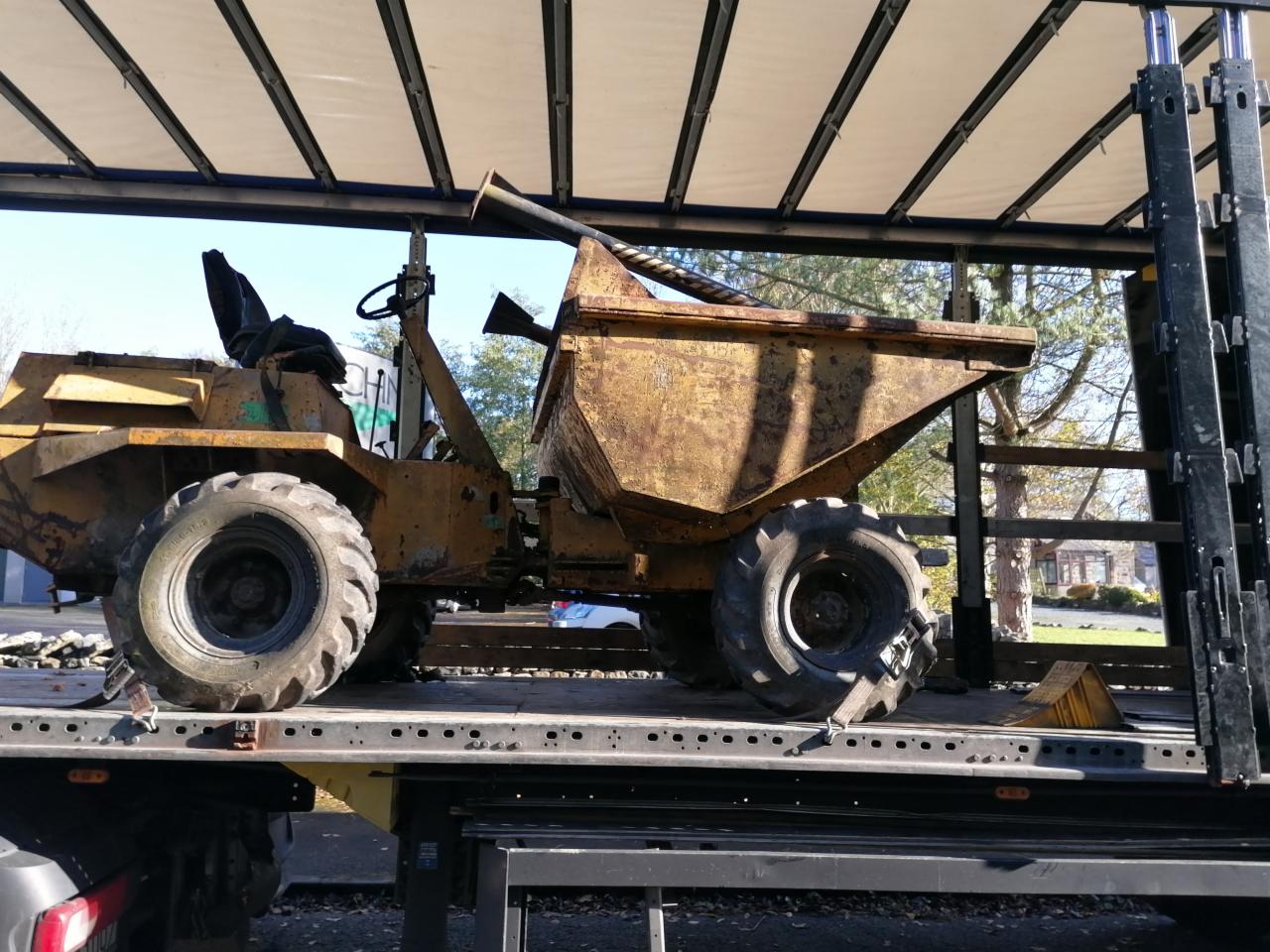 Żółta wywrotka budowlana, z widocznymi śladami użytkowania, zabezpieczona na platformie transportowej pod plandeką, gotowa do przewozu.