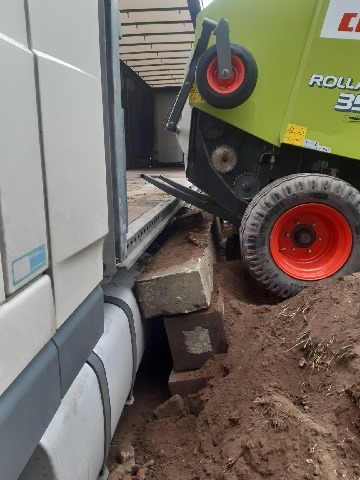 Załadunek prasy rolniczej Claas Rollant 350 do naczepy ciężarówki, zabezpieczonej betonowymi blokami na nierównym terenie.