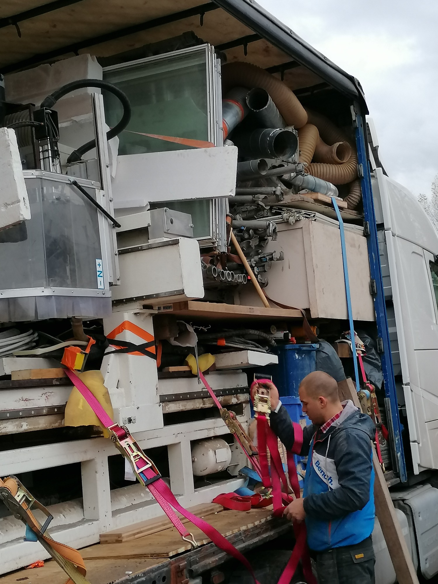 Mężczyzna zabezpiecza ładunek w ciężarówce za pomocą różowych pasów transportowych; w tle maszyny, rury i inne materiały.