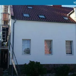 Biała elewacja domu jednorodzinnego z czerwonym dachem, widoczne rusztowanie przy bocznej ścianie budynku.